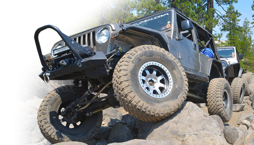 gray Jeep TJ Wrangler crawling over large boulders with black metalcloak fenders and black tow winch, with green pine trees and a white Jeep behind